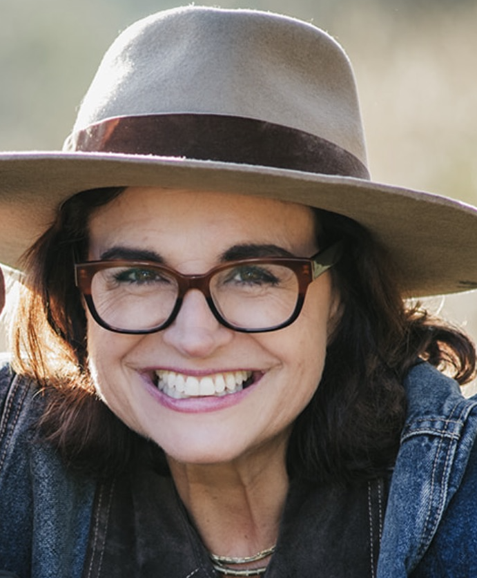 Photo of Lisa Bertelsen-Kivett smiling and wearing a brown hat, brown eyeglasses and blue shirt.